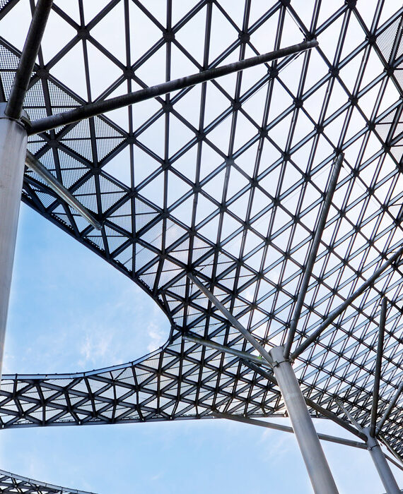 Modern lattice roof structure supported by tall columns against a blue sky.