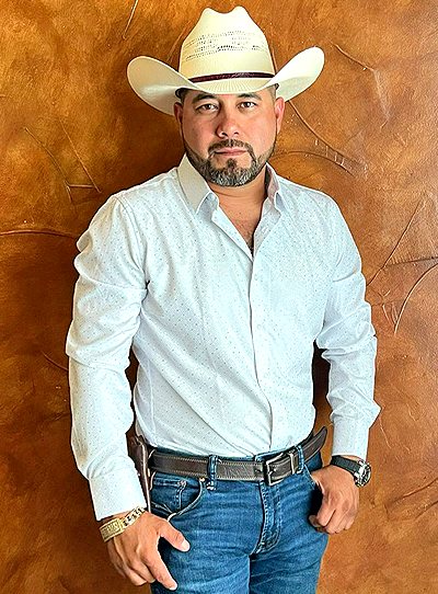 Man in a white shirt and cowboy hat posing against a brown textured wall.